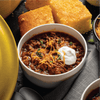 A bowl of chili topped with shredded cheese, sour cream, and chives, with cornbread in the background.
