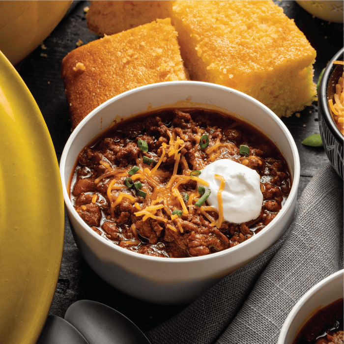 A bowl of chili topped with shredded cheese, sour cream, and chives, with cornbread in the background.