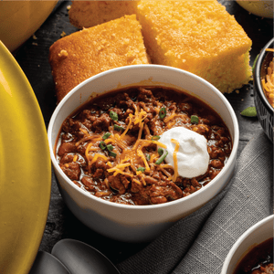 A bowl of chili topped with shredded cheese, sour cream, and chives, with cornbread in the background.