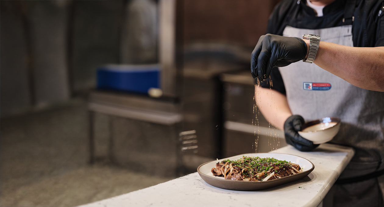 Chef wearing gloves sprinkles seasoning over a plated dish of meat and herbs in a professional kitchen