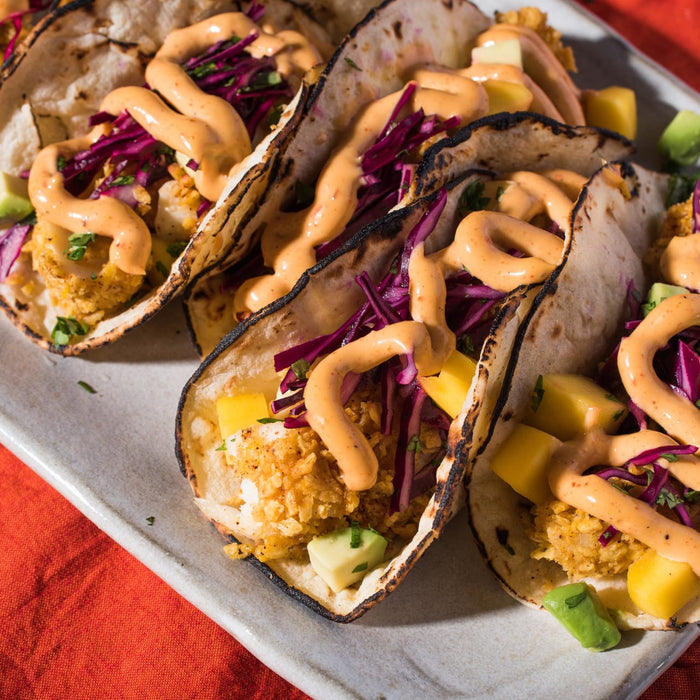 Four tacos filled with fried fish, mango, red cabbage, and avocado, topped with a creamy sauce.