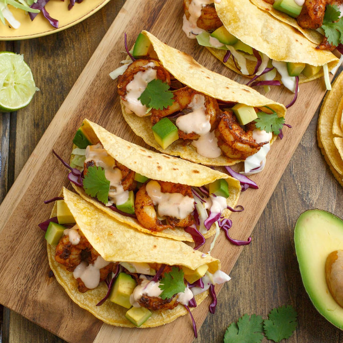 Four shrimp tacos with avocado, cabbage, and cilantro on a wooden cutting board.