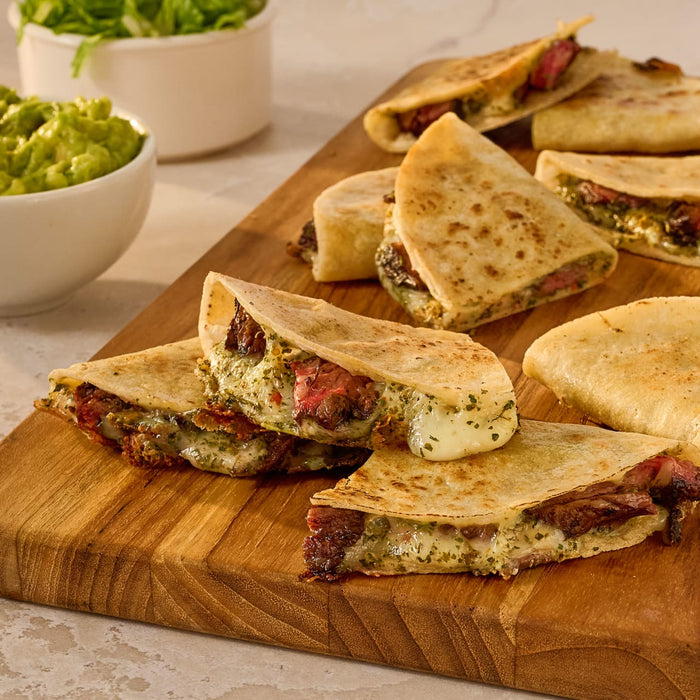 A wooden board displays several quesadillas filled with steak and cheese, accompanied by bowls of guacamole and lettuce.