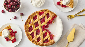 Raspberry pie with slices on plates, whipped cream, and cherries on a white surface.
