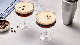 Two espresso martinis with coffee beans on a light gray surface.