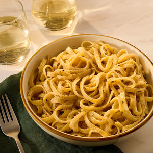 A bowl of fettuccine pasta with herbs and spices is served with two glasses of white wine.