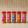A row of five McCormick spice jars, featuring Mccormick Peruvian Style Seasoning 5.48 Oz on the left, alongside Korean, Greek, Indian, and Moroccan style seasonings, all with red caps, on a light countertop against a tiled wall.