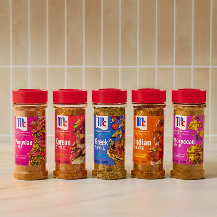 A row of five McCormick spice jars, featuring Mccormick Peruvian Style Seasoning 5.48 Oz on the left, alongside Korean, Greek, Indian, and Moroccan style seasonings, all with red caps, on a light countertop against a tiled wall.