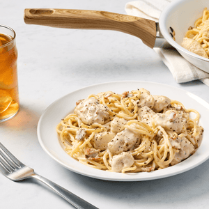A plate of creamy chicken spaghetti, garnished with dried herbs that could be Mccormick Gourmet Oregano Organic, is served alongside a glass of iced tea and a pan of pasta