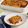 A hearty, cheesy casserole, featuring layers of ground meat, black beans, and corn, and likely prepared using Mccormick Gourmet Paprika, is served on a white plate with a full baking dish in the background, accompanied by lime wedges and jalapeños.