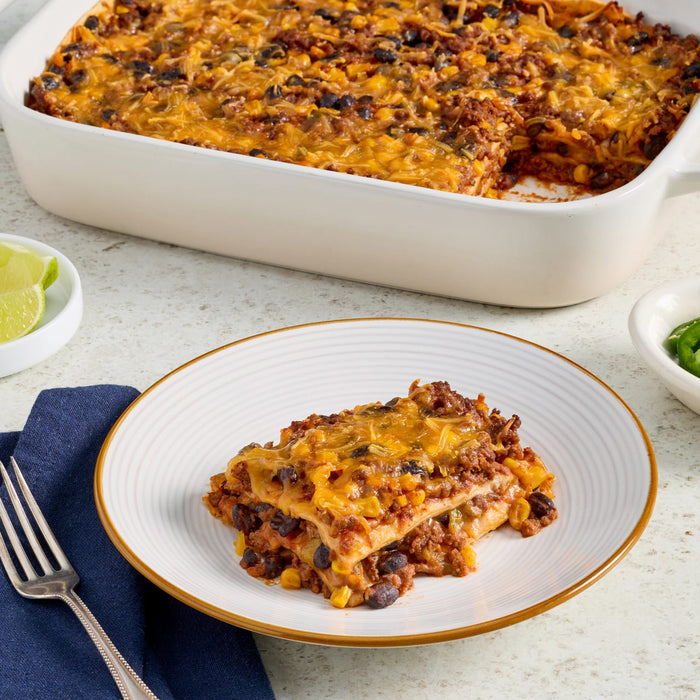 A hearty, cheesy casserole, featuring layers of ground meat, black beans, and corn, and likely prepared using Mccormick Gourmet Paprika, is served on a white plate with a full baking dish in the background, accompanied by lime wedges and jalapeños.