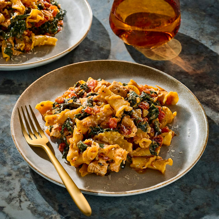 A plate of pasta with a creamy tomato sauce and greens is shown next to a glass of amber liquid, suggesting a delicious and comforting meal.