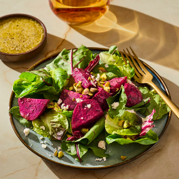 A colorful salad featuring roasted beets, crumbled feta, and chopped pistachios on a bed of mixed greens, a dish that pairs well with Mccormick Gourmet Vp Gmt Basics 1, is presented on a blue plate with a side of dressing and a drink.