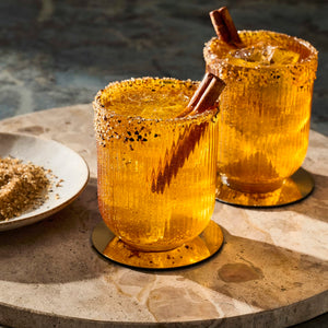 Two cocktails rimmed with sugar and spices, garnished with cinnamon sticks, sit on a stone surface next to a dish of McCormick spices, highlighting a festive and flavorful beverage presentation.