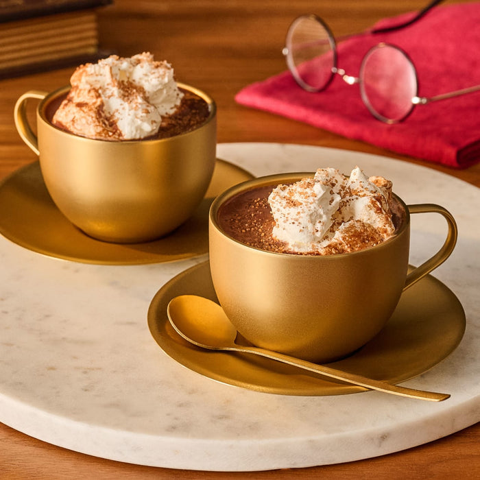 Two golden cups of hot chocolate, topped with whipped cream and a dusting of Mccormick Ravenclaw Finishing Sugar Harry Potter 3.37 Oz, sit on a white marble tray with a golden spoon, a red napkin, and spectacles in the background.