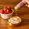 A hand sprinkles Mccormick Slytherin Finishing Sugar Harry Potter 3.37 Oz onto a creamy dessert in a gold bowl, with a bowl of fresh strawberries nearby on a wooden table.