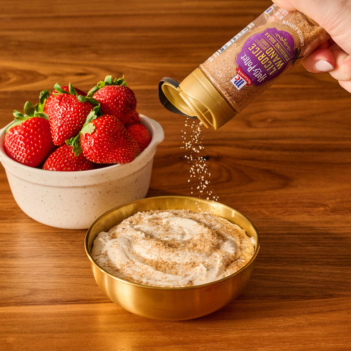 A hand sprinkles Mccormick Slytherin Finishing Sugar Harry Potter 3.37 Oz onto a creamy dessert in a gold bowl, with a bowl of fresh strawberries nearby on a wooden table.