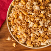 A bowl of seasoned snack mix, featuring popcorn, cereal rings, and other crunchy pieces, rests on a wooden surface beside a red napkin, presented as a delightful treat from the makers of Mccormick Slytherin Finishing Sugar Harry Potter 3.37 Oz.