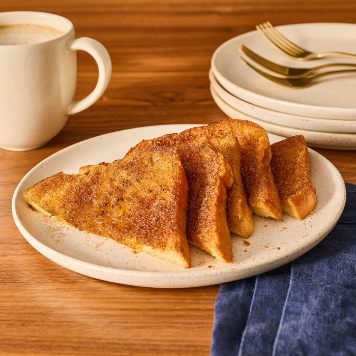 Slices of French toast, generously sprinkled with Mccormick Hufflepuff Finishing Sugar Harry Potter 3.37 Oz, are arranged on a white plate next to a coffee mug and a stack of plates with gold forks.