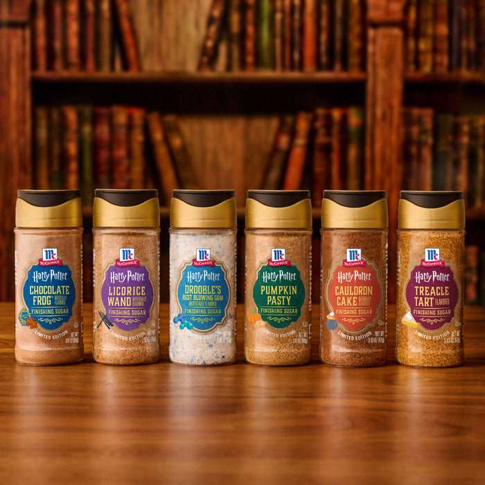 Six McCormick Harry Potter finishing sugars, including the Mccormick Gryffindor Finishing Sugar Harry Potter 3.37 Oz, are arranged on a wooden surface with a blurred bookshelf background.