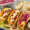Three tacos filled with fish, red cabbage, and cilantro are drizzled with a creamy sauce.