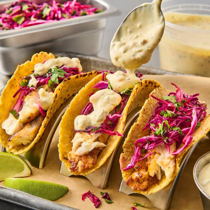 Three tacos filled with fish, red cabbage, and cilantro are drizzled with a creamy sauce.