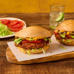 Two crispy breaded sandwiches layered with avocado, tomato, jalapenos, and creamy sauce on a wooden board next to a side salad and a glass of lime water.