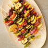 An overhead view of a platter piled with grilled vegetable skewers, featuring zucchini, yellow squash, red onion, and bell peppers, all seasoned with herbs.