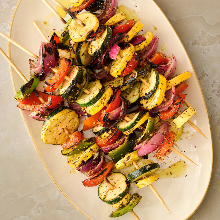 An overhead view of a platter piled with grilled vegetable skewers, featuring zucchini, yellow squash, red onion, and bell peppers, all seasoned with herbs.