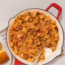 Red skillet with a mixture of food, including sausage and vegetables, on a white surface.