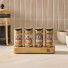 The Mccormick Finishing Sugar Caddy 4Ct is displayed on a kitchen counter, holding jars of Cinnamon Roll, Vanilla Cake, Chocolate Hazelnut, and Salted Caramel finishing sugars next to a French press and coffee mugs.