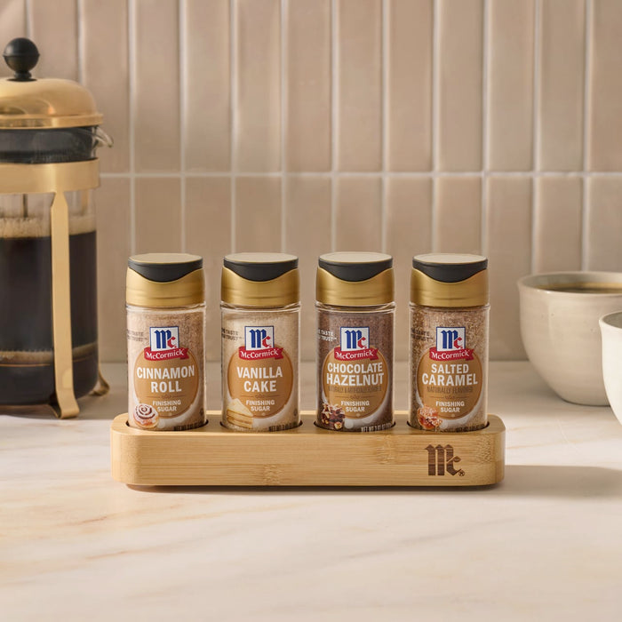 The Mccormick Finishing Sugar Caddy 4Ct is displayed on a kitchen counter, holding jars of Cinnamon Roll, Vanilla Cake, Chocolate Hazelnut, and Salted Caramel finishing sugars next to a French press and coffee mugs.