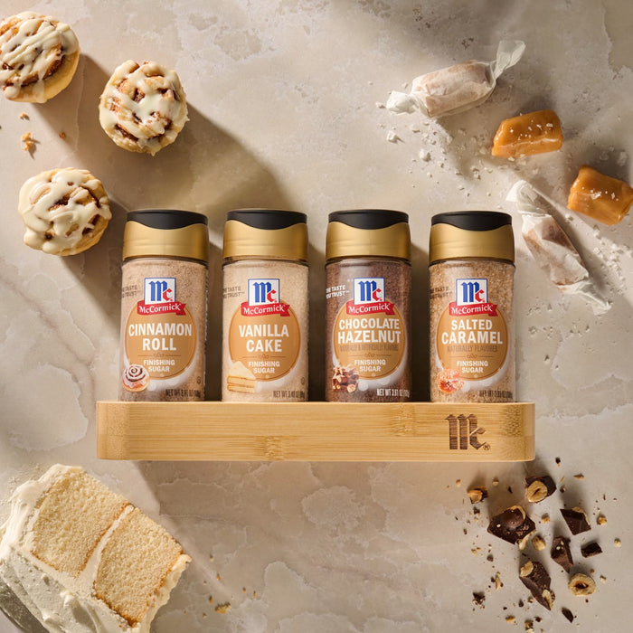 Four McCormick Finishing Sugars are displayed in a wooden holder, part of the Mccormick Finishing Sugar Caddy 4Ct, with each flavor surrounded by its corresponding treat: cinnamon rolls, vanilla cake, chocolate with hazelnuts, and salted caramels.