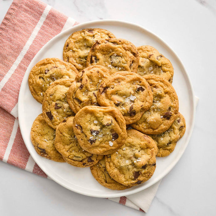 A white plate is filled with golden-brown chocolate chip cookies, generously sprinkled with coarse sea salt, suggesting the use of Mccormick Hss Sea Salt Coarse Ground 13.5 Oz.