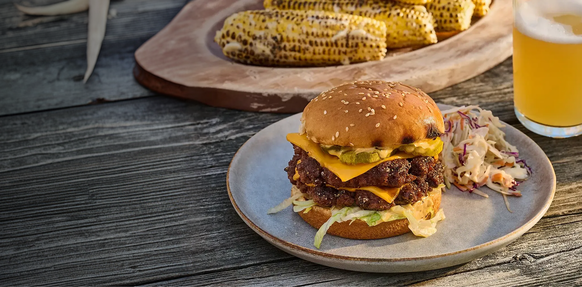 Burger with cheese and lettuce on a plate with corn on the cob and a beer in the background.