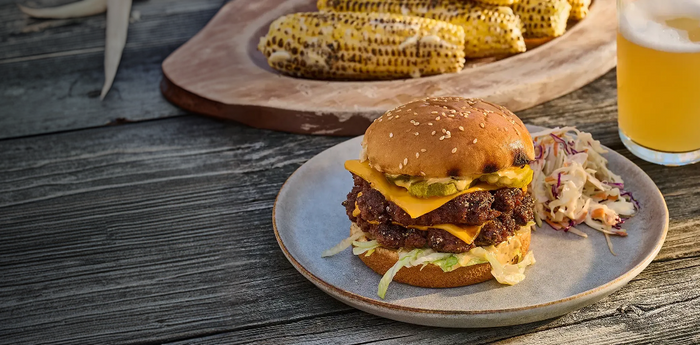 Burger with cheese and lettuce on a plate with corn on the cob and a beer in the background.