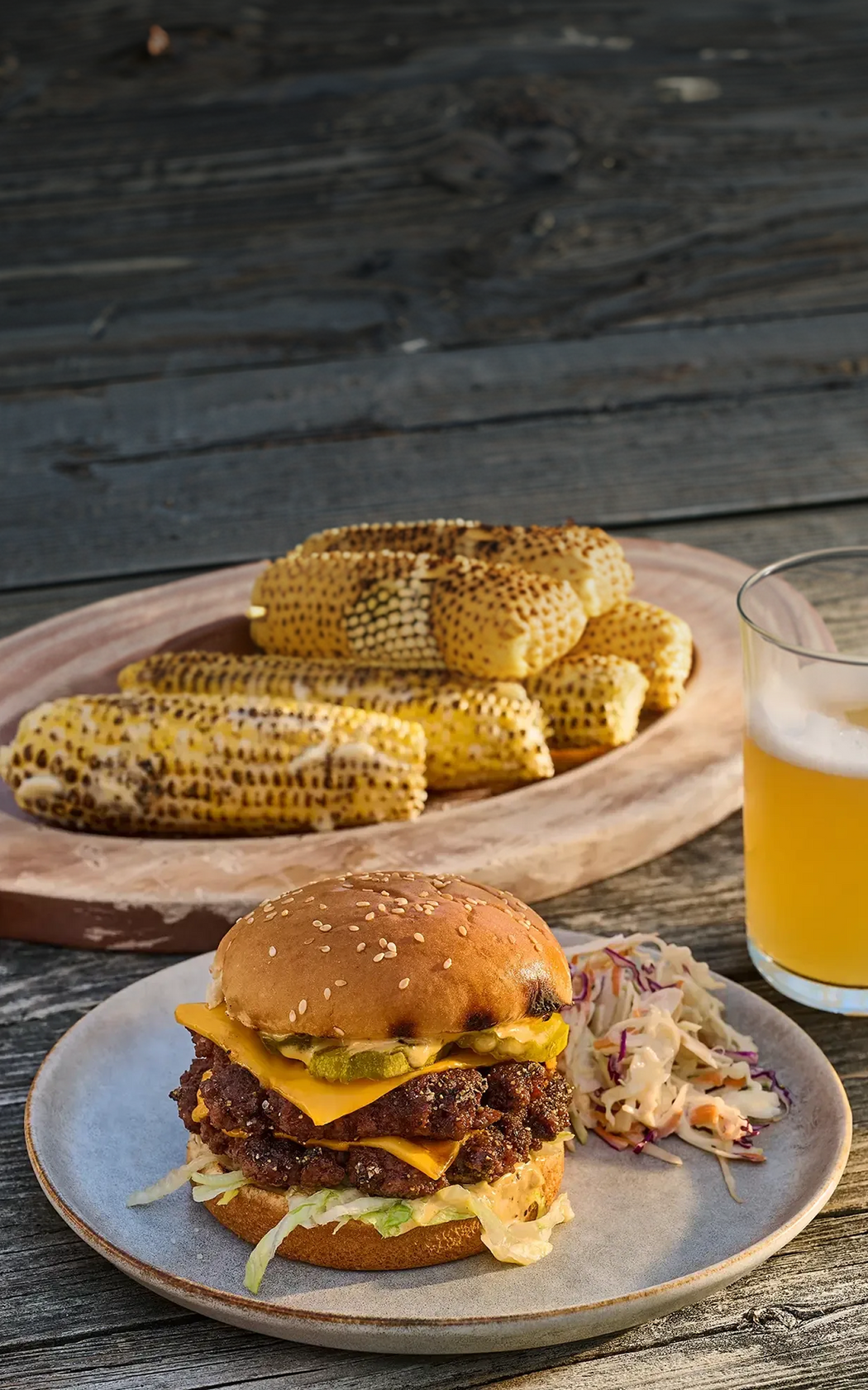 Hamburger with cheese, bacon, and pickles on a plate with corn on the cob and a glass of beer on a wooden surface.