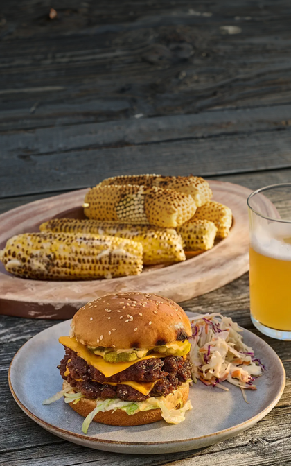 Hamburger with cheese, bacon, and pickles on a plate with corn on the cob and a glass of beer on a wooden surface.