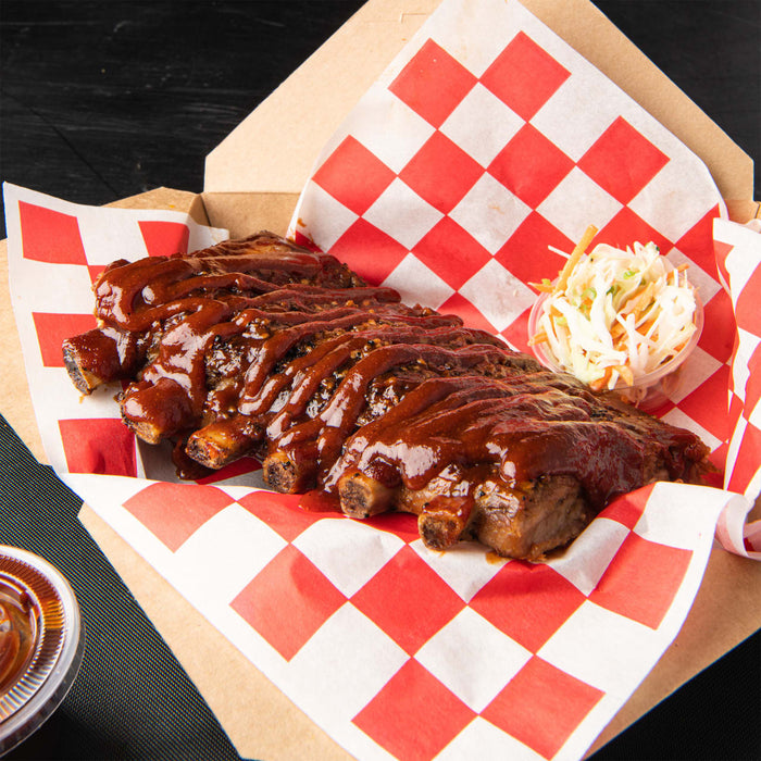 A rack of tender barbecue ribs generously glazed with a rich dark sauce is served in a cardboard takeout container lined with red and white checkered paper alongside a small cup of coleslaw.
