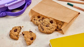Bag of cookies with a few scattered on a surface next to a purple cookie cutter and yellow notebook.