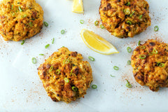 Four crab cakes on a white surface with lemon wedges and green onions.