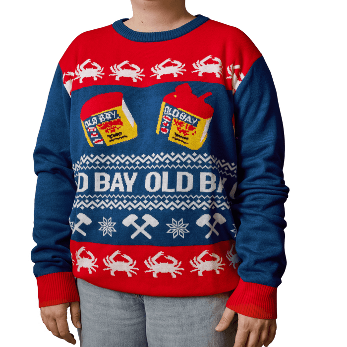 A person wears a festive blue and red holiday sweater featuring the iconic yellow tins and logo of Old Bay seasoning alongside white crab and mallet patterns.