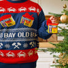 A person wearing a festive holiday sweater featuring crabs and seasoning tins hangs a miniature tin of Old Bay Seasoning as an ornament on a Christmas tree.