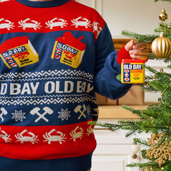 A person wearing a festive holiday sweater featuring crabs and seasoning tins hangs a miniature tin of Old Bay Seasoning as an ornament on a Christmas tree.