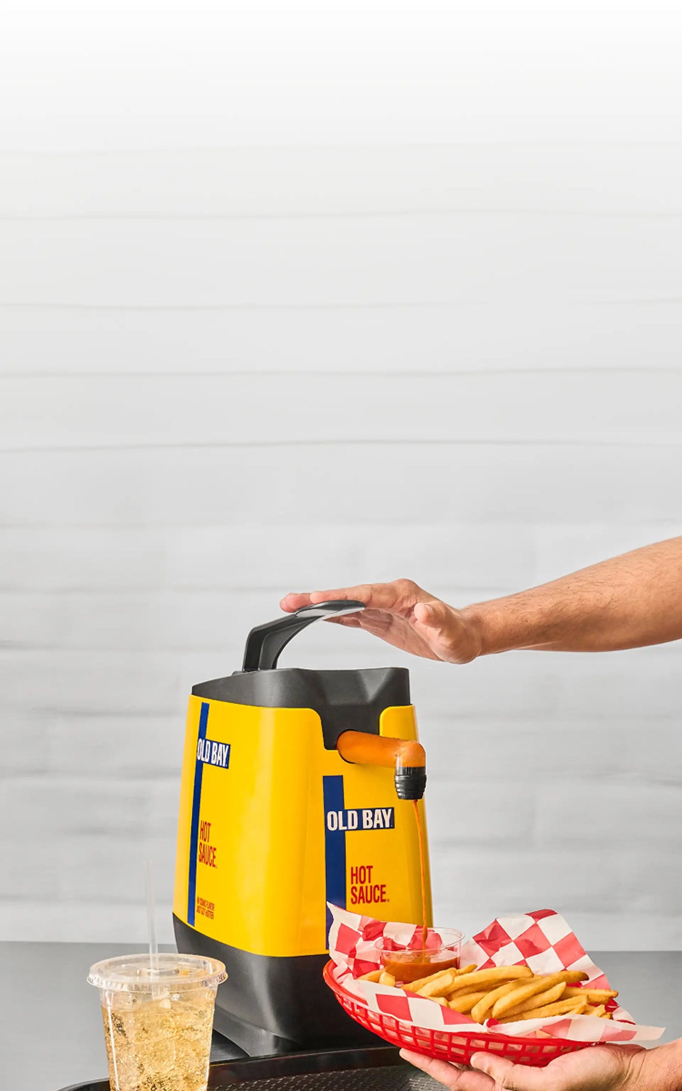Person holding a container of Old Bay hot sauce with a basket of fries on a gray surface.