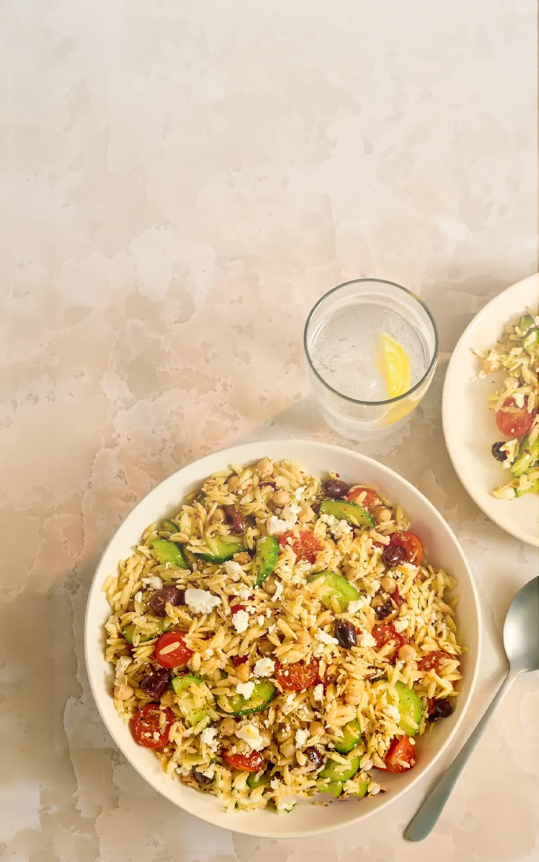 Two bowls of pasta salad with a glass of water and lemon wedge on a textured surface.