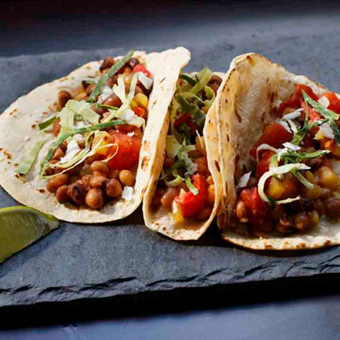 Three tacos filled with black-eyed peas, tomatoes, and cabbage are on a black slate.