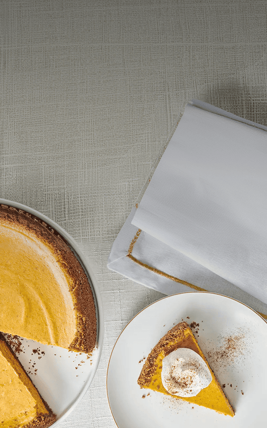 Pumpkin pie with a slice on a plate next to a folded white napkin on a textured surface.