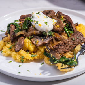 A plate of polenta topped with sauteed mushrooms, spinach, and beef with a dollop of sour cream.