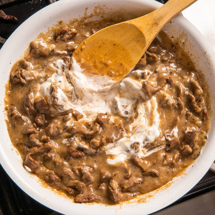 A close-up of a white pan with a wooden spoon in it filled with a creamy beef stew.